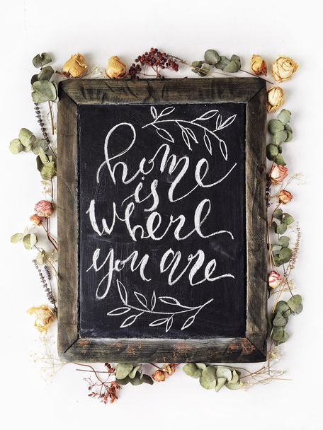 Phrase "Home is where you are" written in calligraphy style at black wooden chalkboard with yellow and pink roses and green leaves - Photo, Image