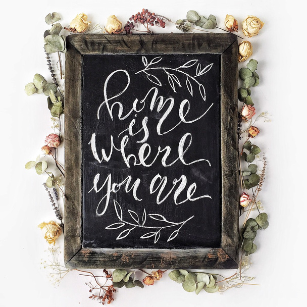 Phrase "Home is where you are" written in calligraphy style at black wooden chalkboard with yellow and pink roses and green leaves - Photo, Image