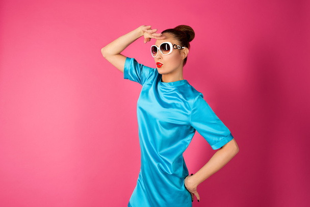 portrait of young beautiful woman in blue silk dress and white sunglasses looking for on the pink background - Photo, Image