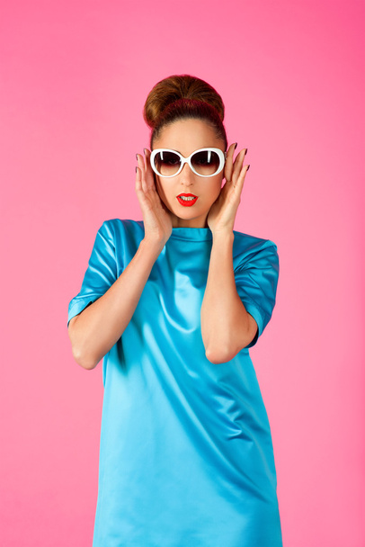 portrait of young smiling beautiful woman in blue silk dress and white sunglasses on the pink background - Foto, imagen