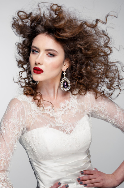 Beautiful young woman with long brown hair, red lips,jewellery in wedding dress Pretty model poses at studio. - Photo, image