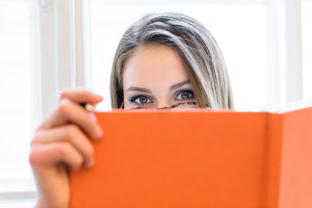 Beautiful blonde student girl covering her face behind the orange book. - Zdjęcie, obraz