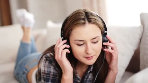 Nuori kaunis nainen makaa sohvalla ja kuuntelee musiikkia hänen kuulokkeet olohuoneessa
. - Materiaali, video