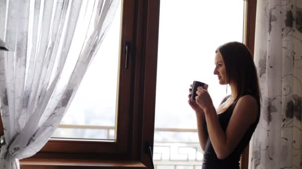 Привлекательная молодая леди отдыхает и пьет чашку чая или кофе перед окном дома
. - Кадры, видео