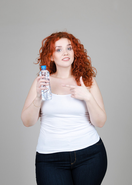 beautiful fat girl with red hair with a bottle of mineral water hands on a gray background - Fotografie, Obrázek