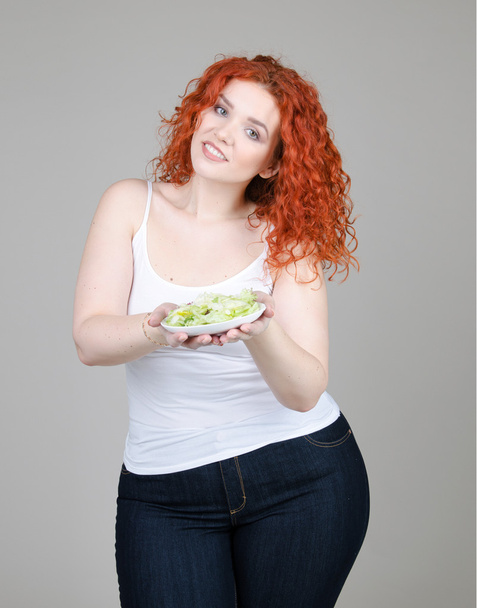 beautiful fat girl with red hair with a plate of salad in hand on gray background - Fotoğraf, Görsel