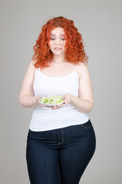 beautiful fat girl with red hair with a plate of salad in hand on gray background - Foto, Imagen