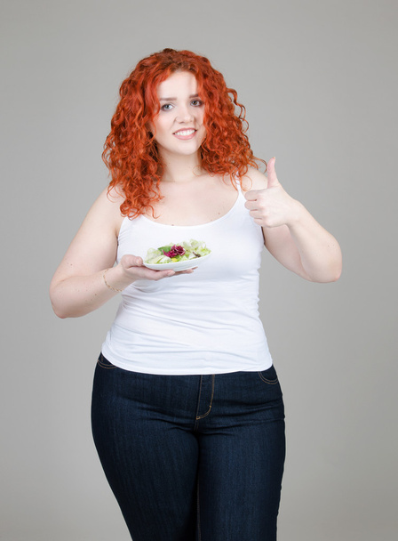 beautiful fat girl with red hair with a plate of salad in hand on gray background - 写真・画像