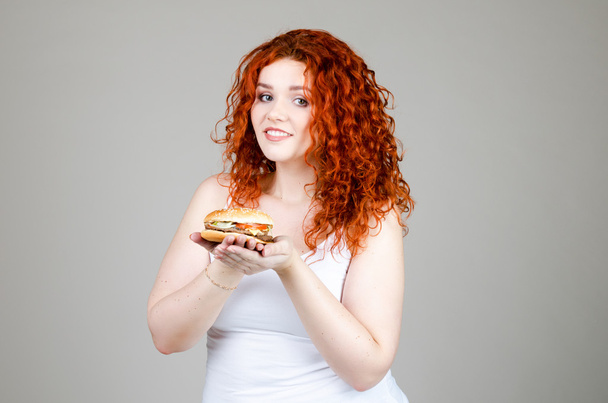 mooie dikke meid met rood haar met hamburger in de hand op grijze achtergrond - Foto, afbeelding