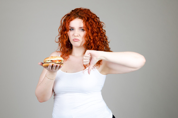 όμορφο χοντρό κορίτσι με κόκκινα μαλλιά με Burger στο χέρι σε γκρι φόντο - Φωτογραφία, εικόνα