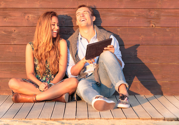 Cheerful guy with girl laugh about video on the tablet. Best friends having fun at the beach with social media and funny contents. Friendship of young people during holiday life moment at sunset. - Photo, Image