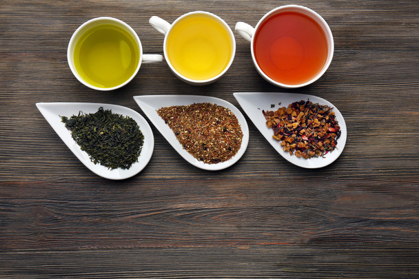 Tea concept. Different kinds of dry tea in ceramic bowls and cups of aromatic tea on wooden background - Photo, image