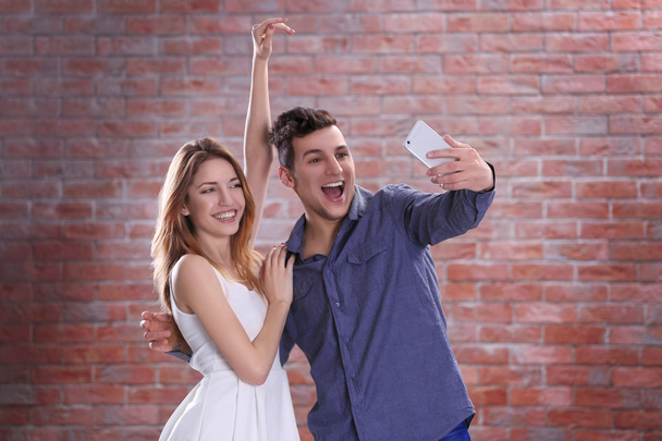 Jovem casal atraente tomando selfie com telefone celular no fundo da parede de tijolo
 - Foto, Imagem