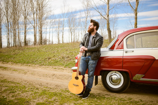Бородатий чоловік грає на гітарі на відкритому повітрі біля ретро-автомобіля
 - Фото, зображення