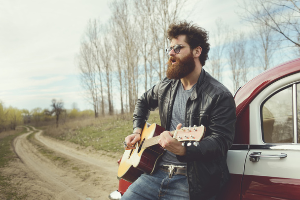 Vousatý muž hrát na kytaru venku poblíž retro auta - Fotografie, Obrázek