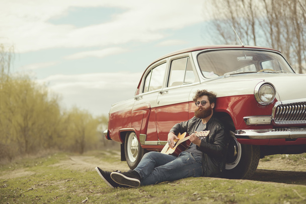 Parrakas mies soittaa kitaraa ulkona lähellä retro auto
 - Valokuva, kuva