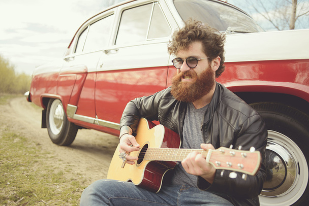 Parrakas mies soittaa kitaraa ulkona lähellä retro auto
 - Valokuva, kuva