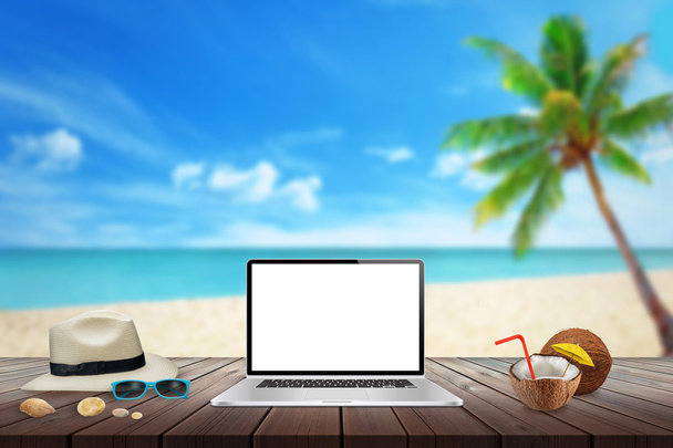 Exibição isolada de laptop na mesa de madeira para maquete. Coco, chapéu, conchas, pedras, óculos de sol na mesa. Praia, mar, palma e céu azul no fundo
. - Foto, Imagem