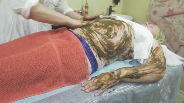 Cosmetologist smears seaweed mixture on fat woman stomach, left hand in saloon - Video, Çekim