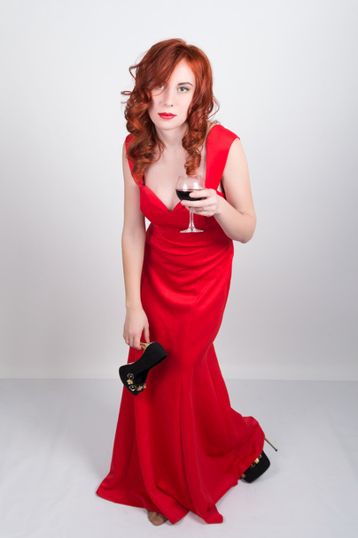 Beautiful young sexy slim red-haired girl wearing a slinky silk red dress high heels, in alcoholic intoxication. drinking red wine from a glass. after party - Photo, image