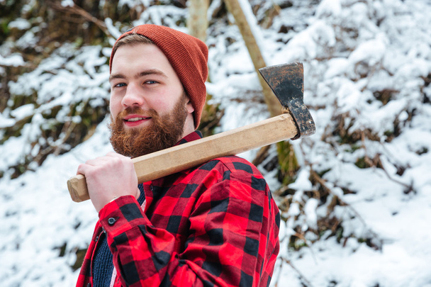 Smiling attractive bearded man with axe in winter forest - Фото, изображение
