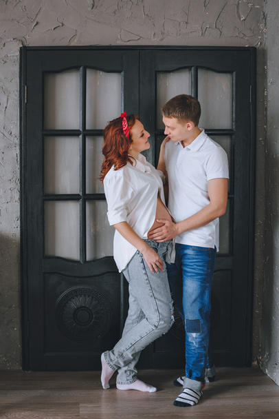 A successful young man and a young beautiful pregnant woman. In anticipation of the baby.  We look at each other and hug the midsection. Photo shoot in the Studio. A woman in jeans and a white shirt. - Photo, Image