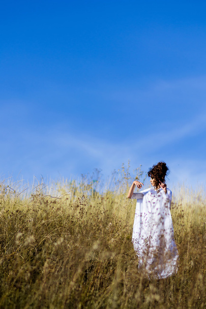 Beautiful,graceful,posing girl stand in field.Goddess, graceful girl in the field with blue sky,white dress, meadow,sunny field. Exquisite, stunning,striking girl poses, raises hands up in the field with flowers, dry grass, cones, grain and looks up. - Foto, Imagem