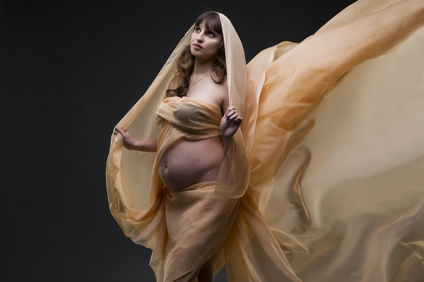 Young beauty and sexy caucasian pregnant woman with yellow silk on gray studio background - Fotó, kép