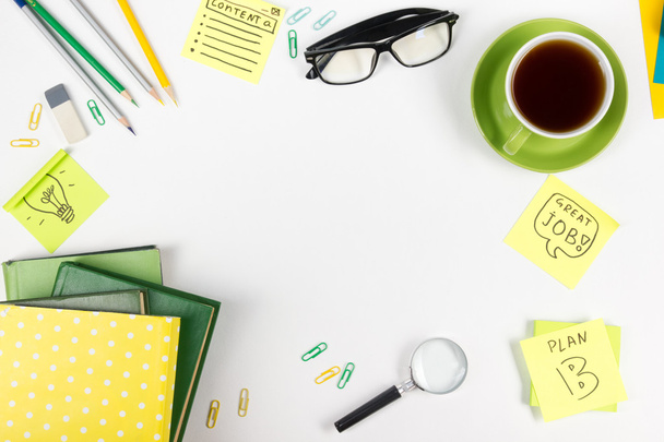Офисный стол с зелеными принадлежностями, блокнот, чашка, ручка, очки, смятая бумага, увеличительное стекло, цветок на белом фоне. Вид сверху
 - Фото, изображение