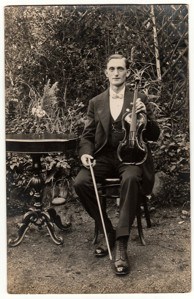 Vintage φωτογραφία δείχνει πόζες νεαρός άνδρας με το βιολί σε εξωτερικούς χώρους. Ο βιολονίστας κάθεται στο μαύρο κομψό τραπέζι. Αντικέ μαύρο & άσπρο φωτογραφία. - Φωτογραφία, εικόνα