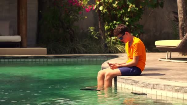teenager sitting with smartphone at the edge of swimming pool - Materiał filmowy, wideo