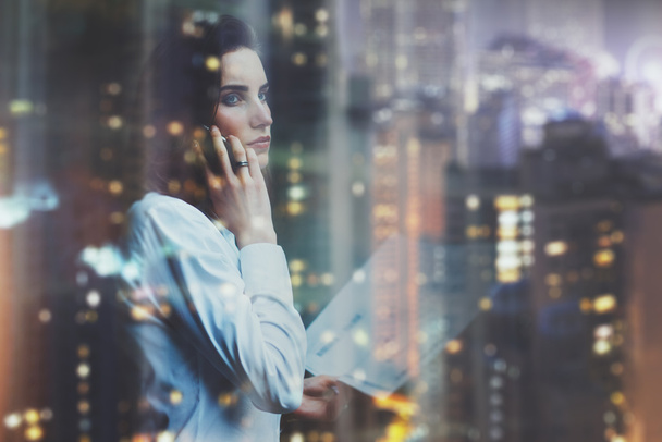 Photo woman wearing white shirt, talking smartphone and holding business documents in hands. Open space loft office. Panoramic windows, night city background. Horizontal mockup, bokeh. Film effect - Φωτογραφία, εικόνα