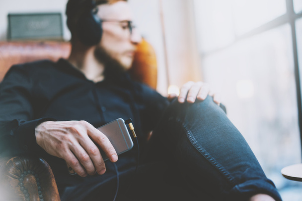 Photo handsome bearded man headphones listening to music modern loft studio.Man sitting in vintage chair looking window,holding smartphone and relaxing.Blurred background.Horizontal, film effect. - 写真・画像