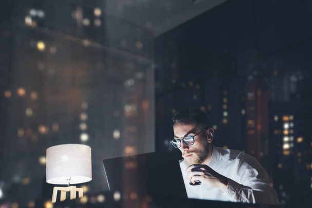 Bearded young businessman working on modern loft office at night. Man using contemporary notebook texting message, holding cup espresso, blurred background. Horizontal, film effect, bokeh - Fotó, kép