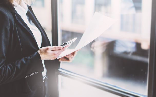 Photo business woman wearing suit, touching smartphone screen and holding documents in hands. Open space loft office. Panoramic windows background. Wide mockup. Film effect - Foto, imagen