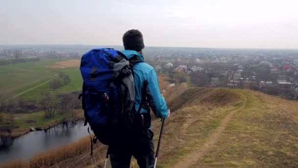 Man tourist in a blue jacket with a backpack and trekking poles walks outdoor - Séquence, vidéo