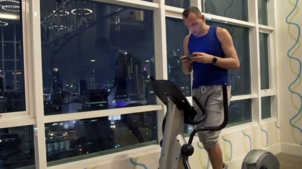 man with smartphone exercising on elliptical machine  - 映像、動画