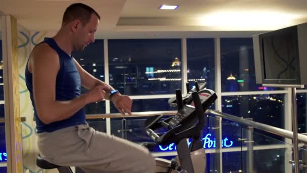 man with smartwatch riding stationary bike in gym - Materiał filmowy, wideo