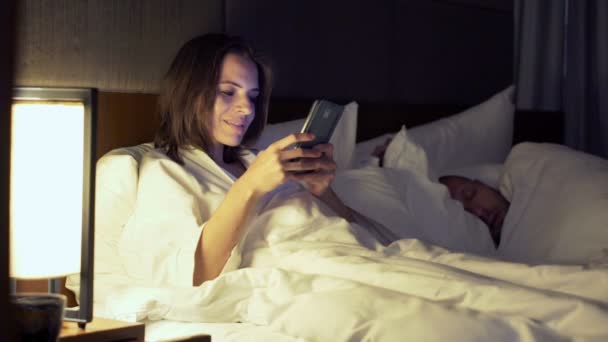woman texting on smartphone and her husband sleeping - Materiał filmowy, wideo