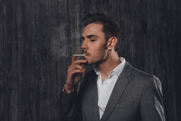 Portrait of young businessman smoking a cigarette on gray backgr - Zdjęcie, obraz