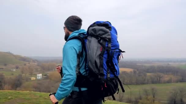 Man tourist in a blue jacket with a backpack and trekking poles walks outdoor - Filmati, video