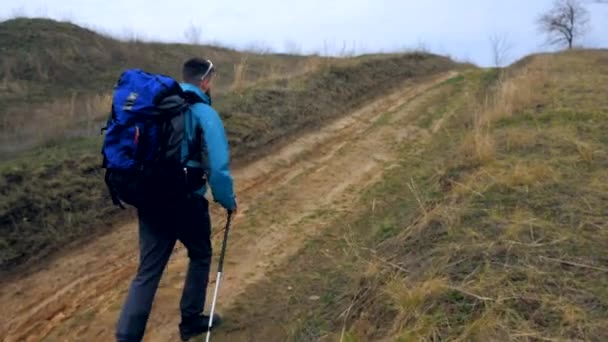 Man tourist in a blue jacket with a backpack and trekking poles walks outdoor - Video