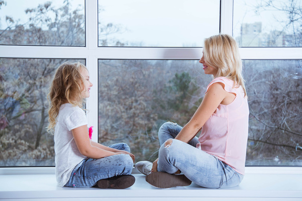 Mom and daughter blonde caucasians are sitting on a windowsill of a large window looking at each other. Beautiful smile. Happy beautiful family. Mothers Day - Фото, изображение