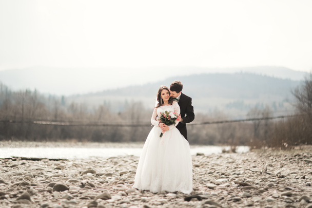 Beautifull wedding couple kissing and embracing near river with stones - Фото, изображение