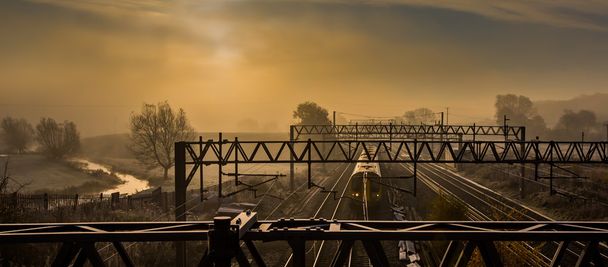 Mattina treno espresso che passa lungo i binari in una mattina nebbiosa, con il fiume bagnato da un bagliore dorato dall'alba
 - Foto, immagini