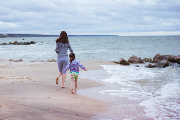 Young european  mother and her little daughter walking along the sandy coast of the sea, playing in the waves, wet feet in the sea, running along the coast. Cloudy and windy weather at sea - Photo, Image