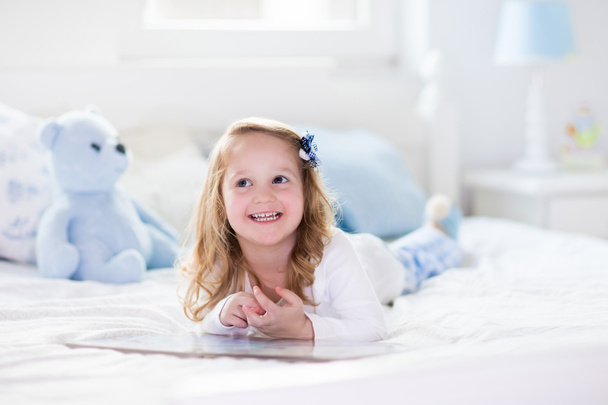 Маленькая девочка играет с игрушкой и читает книгу в постели
 - Фото, изображение