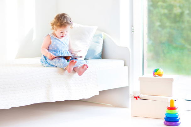 Urocza dziewczyna malucha czytając książkę siedzącą na białym łóżku - Zdjęcie, obraz