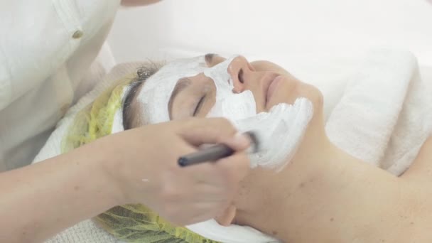 Cosmetólogo poner máscara de arcilla blanca en la cara de la mujer por el cepillo en salón de belleza
 - Metraje, vídeo