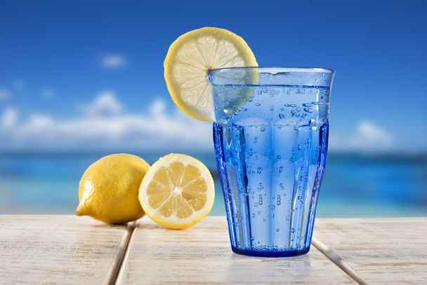 A Blue glass with sparkling water and lemon on a wooden deck overlooking a tropical beach - focus on the lemon - Photo, Image